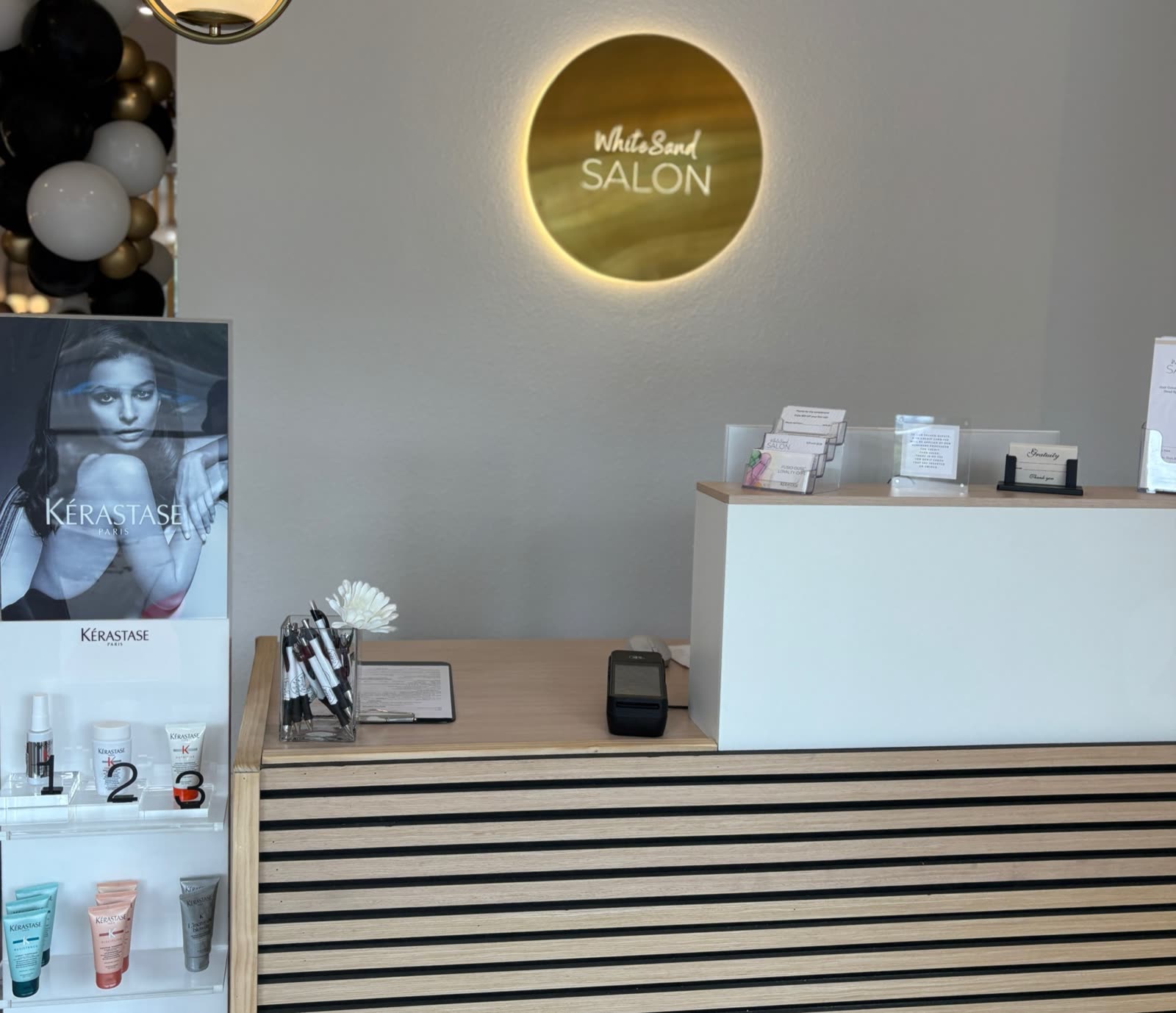 The reception desk at White Sand Salon in Clearwater, the Kérastase home-care display standing beside the backlit gold salon sign